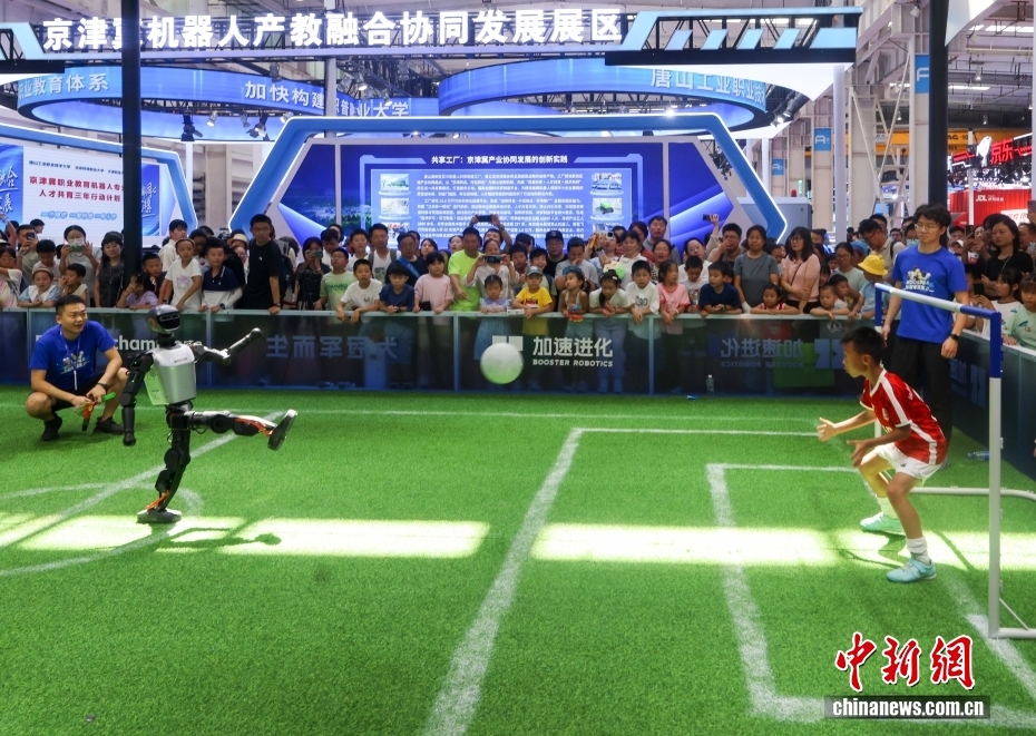 中新社记者 贾天勇 摄 8月11日，2025世界机器人大会继续在北京亦庄举行，展出的各类机器人刮起运动旋风。图为人形机器人展示足球射门技能，一名小朋友上场担任守门员。